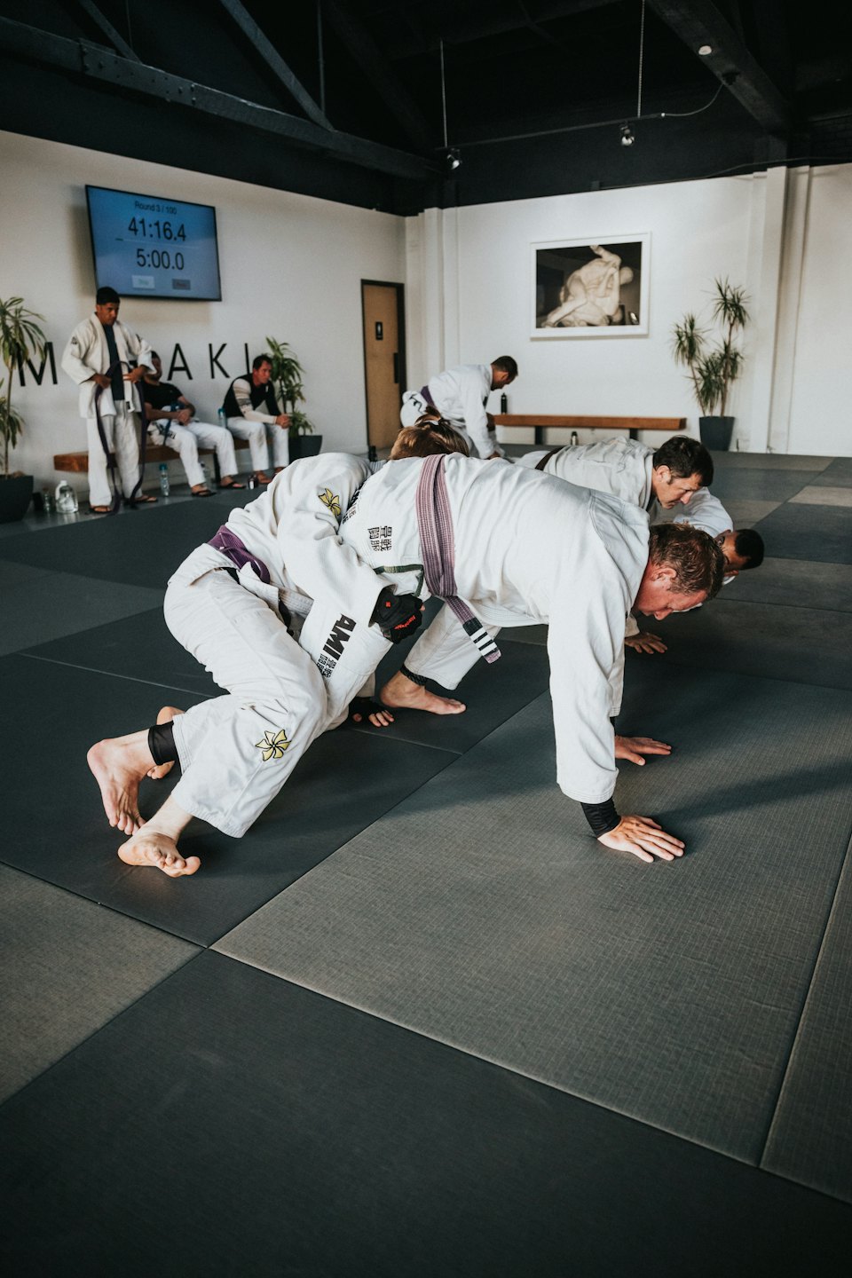 Uomini in gi bianchi fanno flessioni di Jiu-Jitsu brasiliano su tappetini grigi.